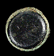 Copper alloy button back with a dark black or purple glass inset, flat on the front and beveled at the edge. The copper corrosion of the button back is toxic to microorganisms, which likely explains how some fabric that was in contact with the button survived and is still attached to the button back. The weave is visible, and the fabric is probably linen.
